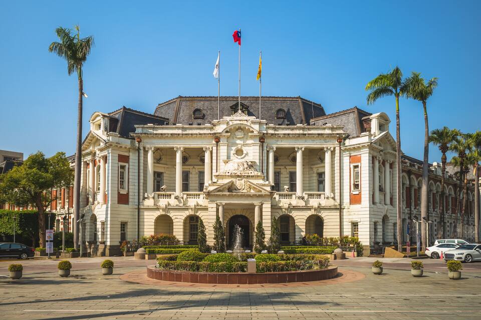 Un imponente edificio neoclásico con una fuente central, rodeado de palmeras y un jardín cuidado.