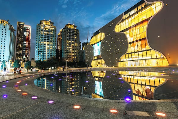 A modern cityscape with illuminated buildings and a reflective water feature.