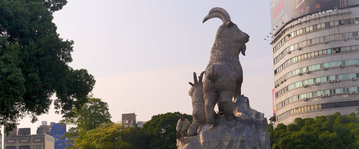 A large metal sculpture of a ram in a park with trees and buildings in the background.