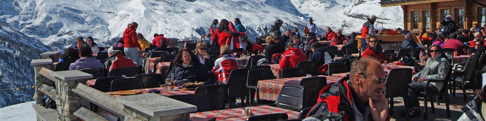 Un paysage montagneux enneigé, avec un sommet imposant, un espace repas avec des tables et des chaises, et des personnes réunies autour.