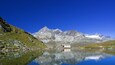 Un lago de montaña con una cabaña pequeña y terreno rocoso.