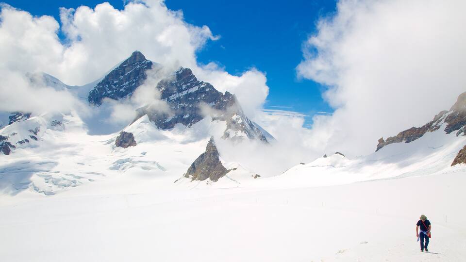 A person hiking on a snow-covered mountain with rocky peaks and clouds in the background.