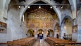 An interior of a church with wooden pews, a large mural on the wall, and a man taking a photo.