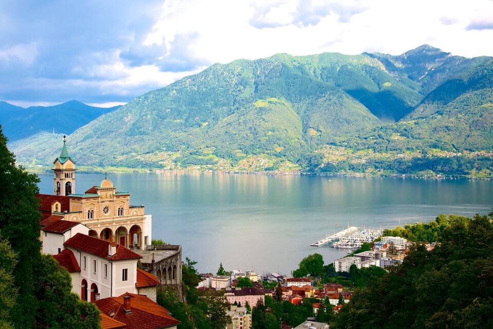 Ein See, umgeben von Bergen, eine Kirche mit rotem Dach und ein Hafen mit Booten.