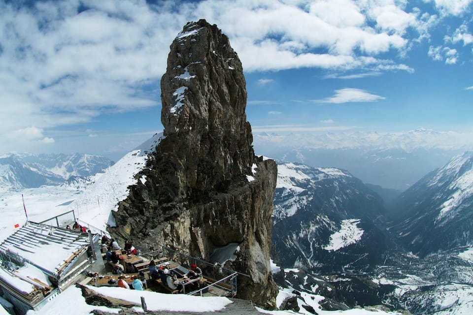 Cumbre nevada de montaña con una plataforma de observación y gente abajo.