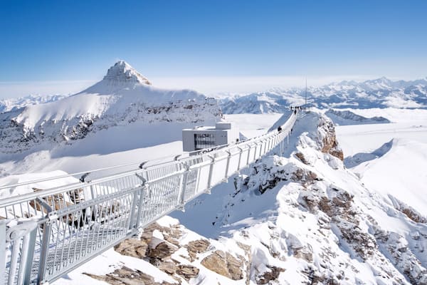 Ein schneebedeckter Berggipfel mit einer Aussichtsplattform und Metallgeländer, mit Blick auf eine weite, verschneite Landschaft.