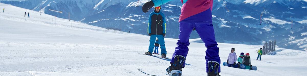 Ein Snowboarder auf einer grünen Schiene in einem Skigebiet, im Hintergrund verschneite Berge.