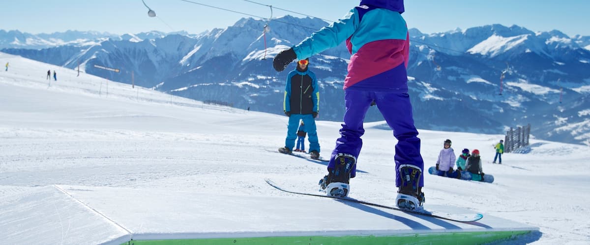 A snowboarder on a green rail at a ski resort with snow-covered mountains in the background.
