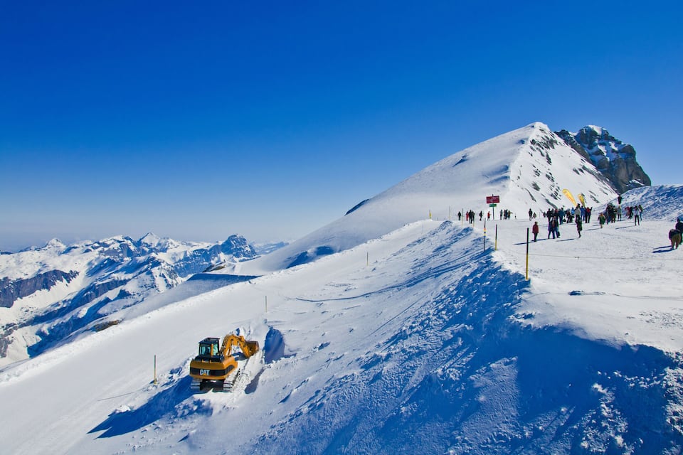 A snow-covered mountain with a yellow excavator and a crowd of people on a slope.