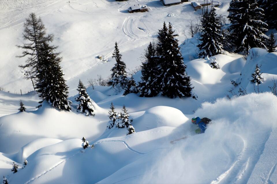 Un paysage de montagne enneigé, avec des traces de ski, des arbres à feuilles persistantes et un chalet en arrière-plan.