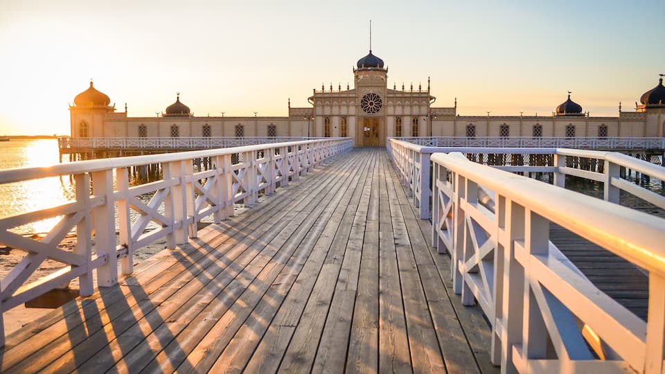 A wooden pier leading to a large building with domed roofs and a clock tower.