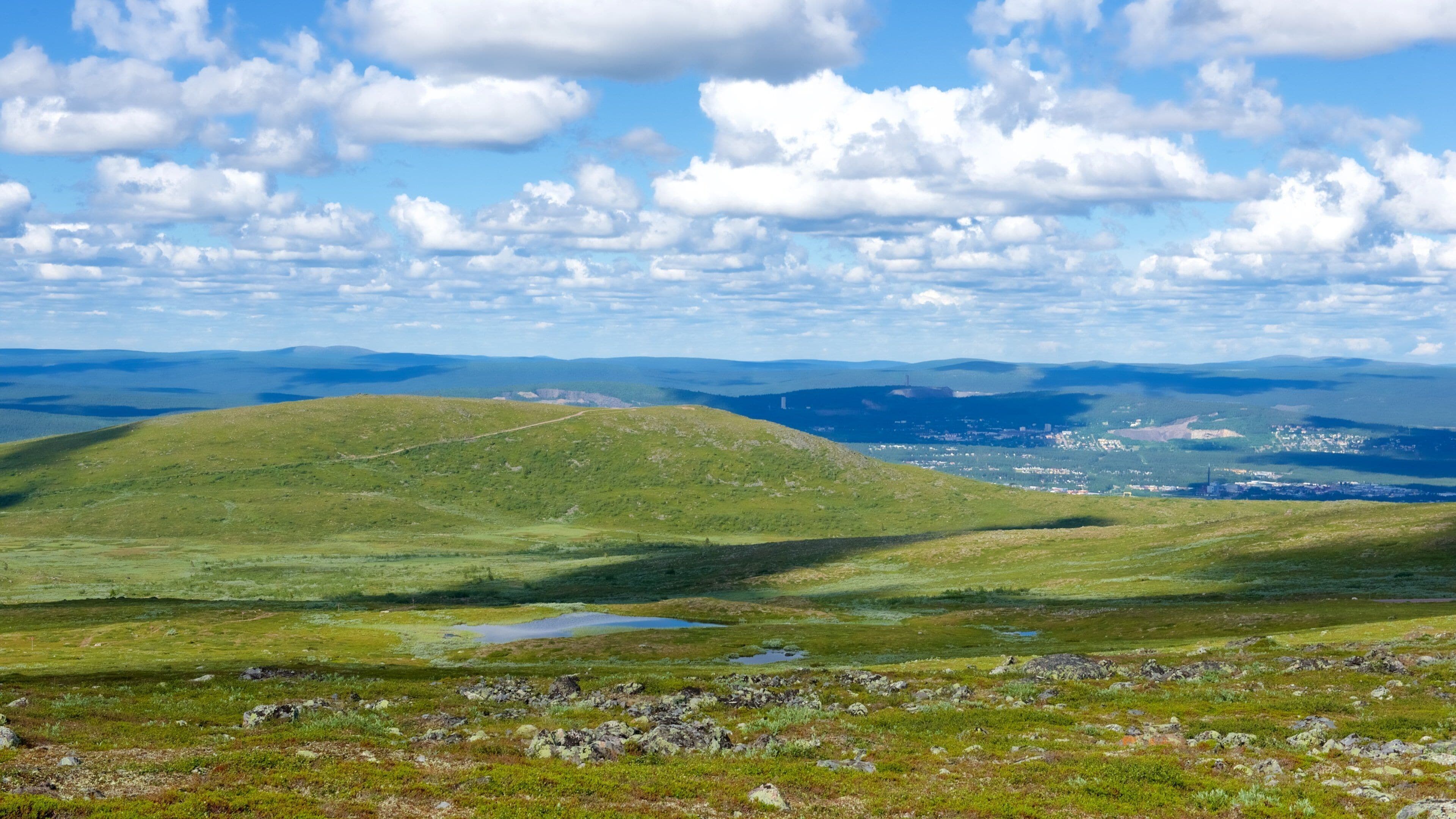 Eine gebirgige Landschaft mit sanften Hügeln, einem kleinen See und einer entfernten Stadtansicht.