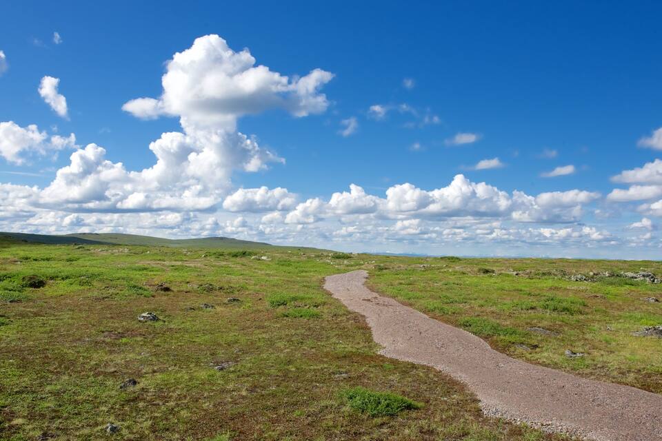 Uma trilha de cascalho serpenteando por um campo gramado sob um céu azul com nuvens dispersas.