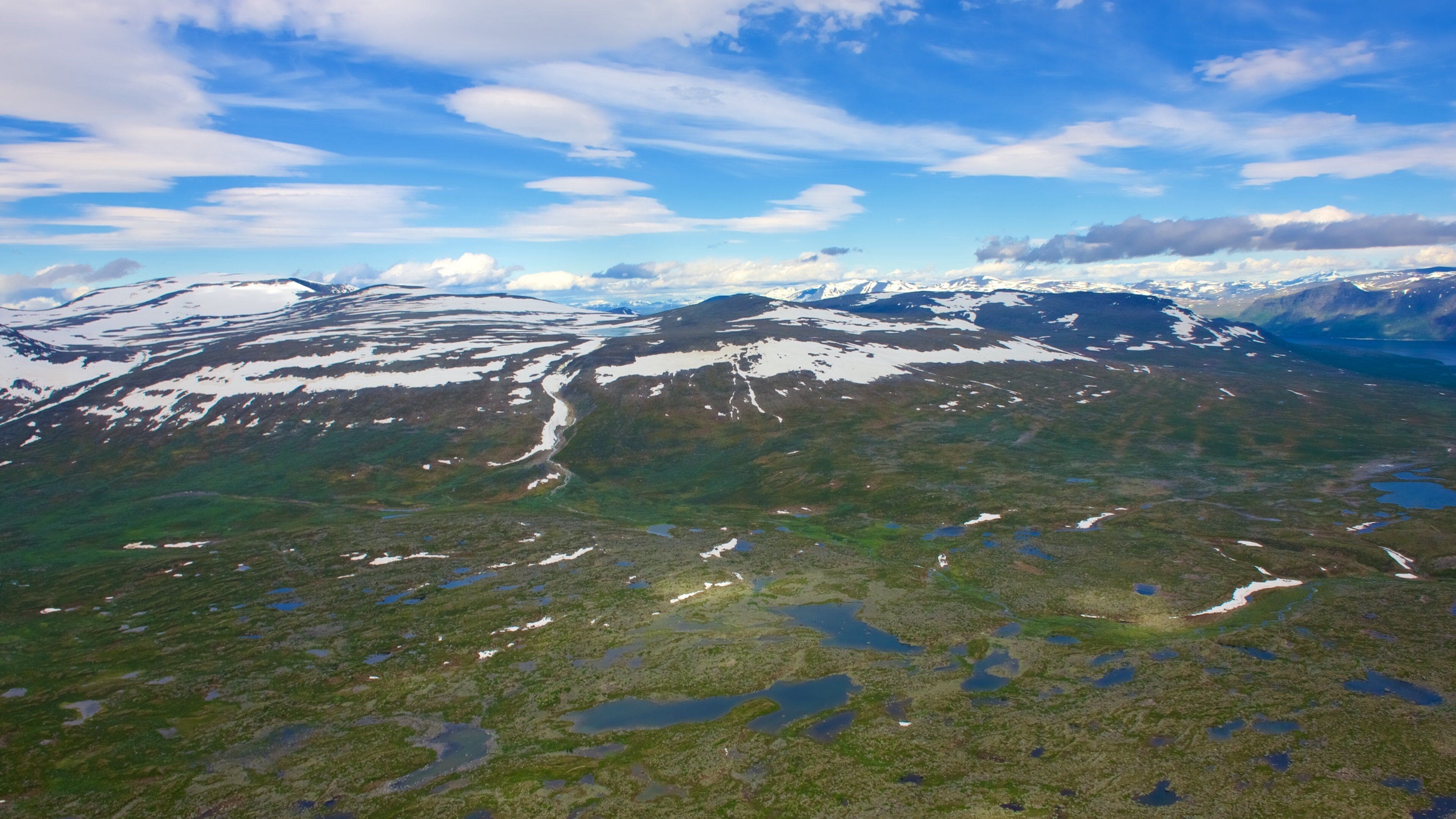 A mountainous landscape with snow-capped peaks, green valleys, and scattered lakes.