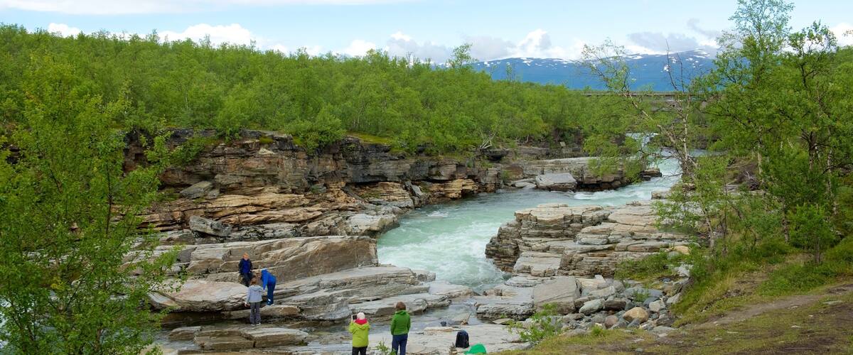 A rocky river gorge with people standing on the rocks, surrounded by green trees and mountains in the background.