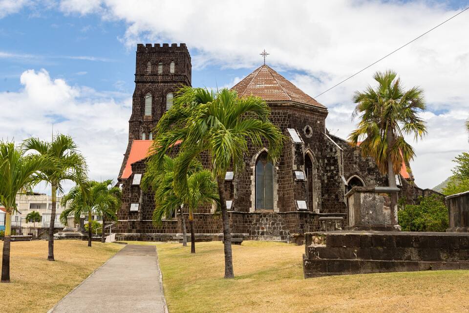 A historic church with a tall tower and a red-tiled roof, surrounded by palm trees and a paved walkway.