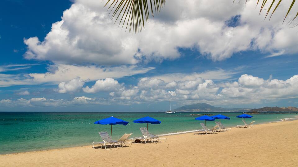 A beach with clear turquoise water, a sailboat in the distance, and a mountain range on the horizon.