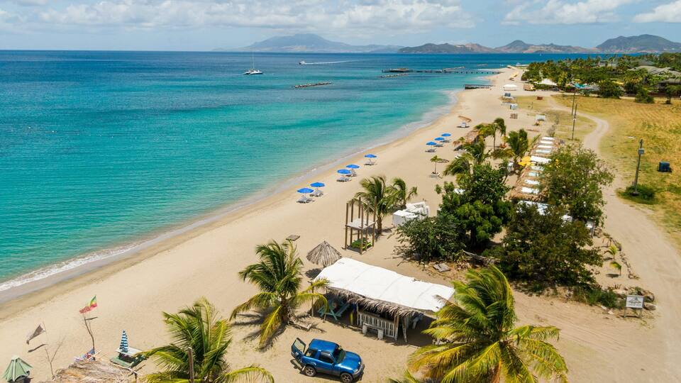 A beach with clear turquoise waters, a row of beach umbrellas, and a few boats in the distance.