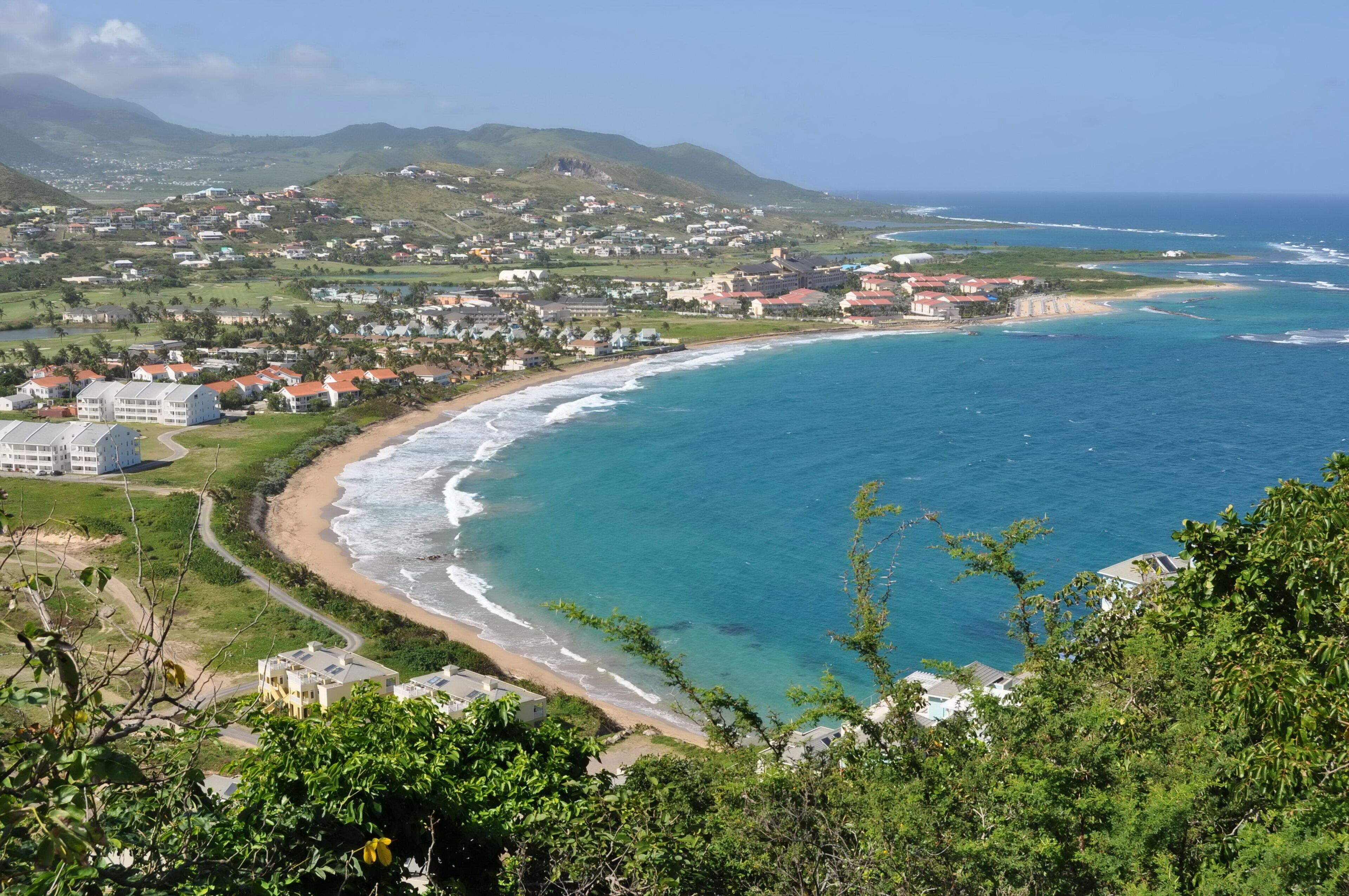 Uma vila costeira com praia, oceano e colinas ao fundo.
