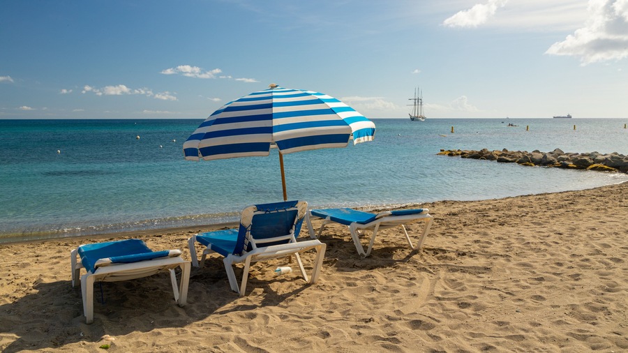 Ein Strand mit einem gestreiften Sonnenschirm, zwei blauen Liegestühlen und einem Felssteg im Hintergrund.