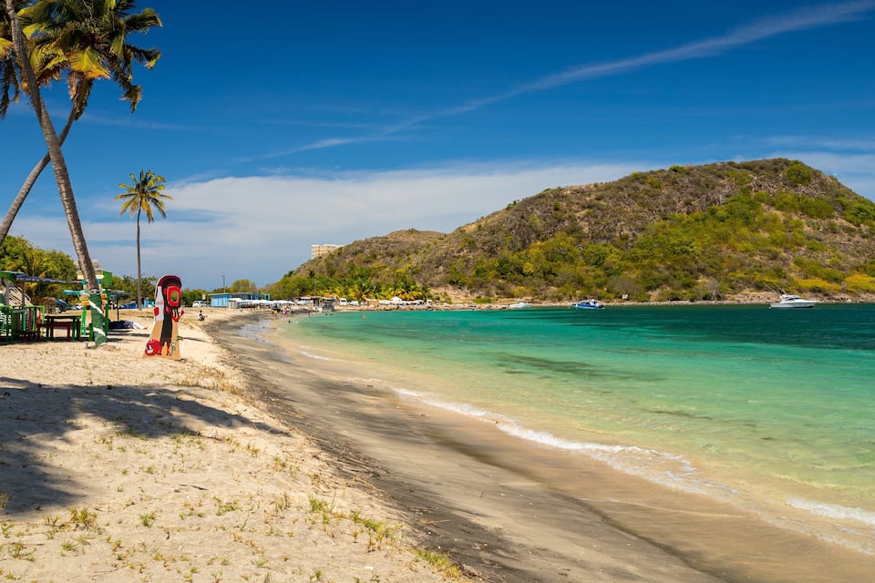 A beach with clear turquoise water, palm trees, and a hilly landscape in the background.