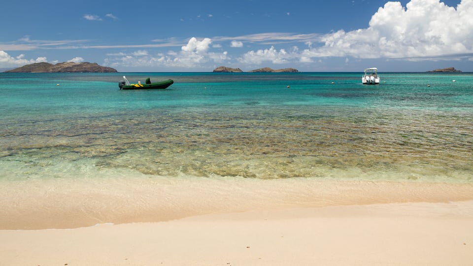 Una playa con agua turquesa cristalina, una orilla arenosa y dos barcos a lo lejos.