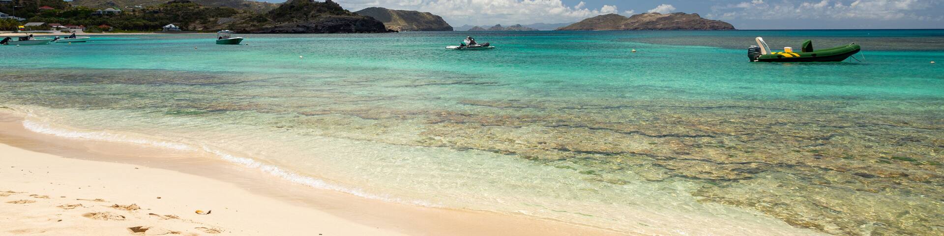 A beach with clear turquoise water, footprints in the sand, and boats in the distance.