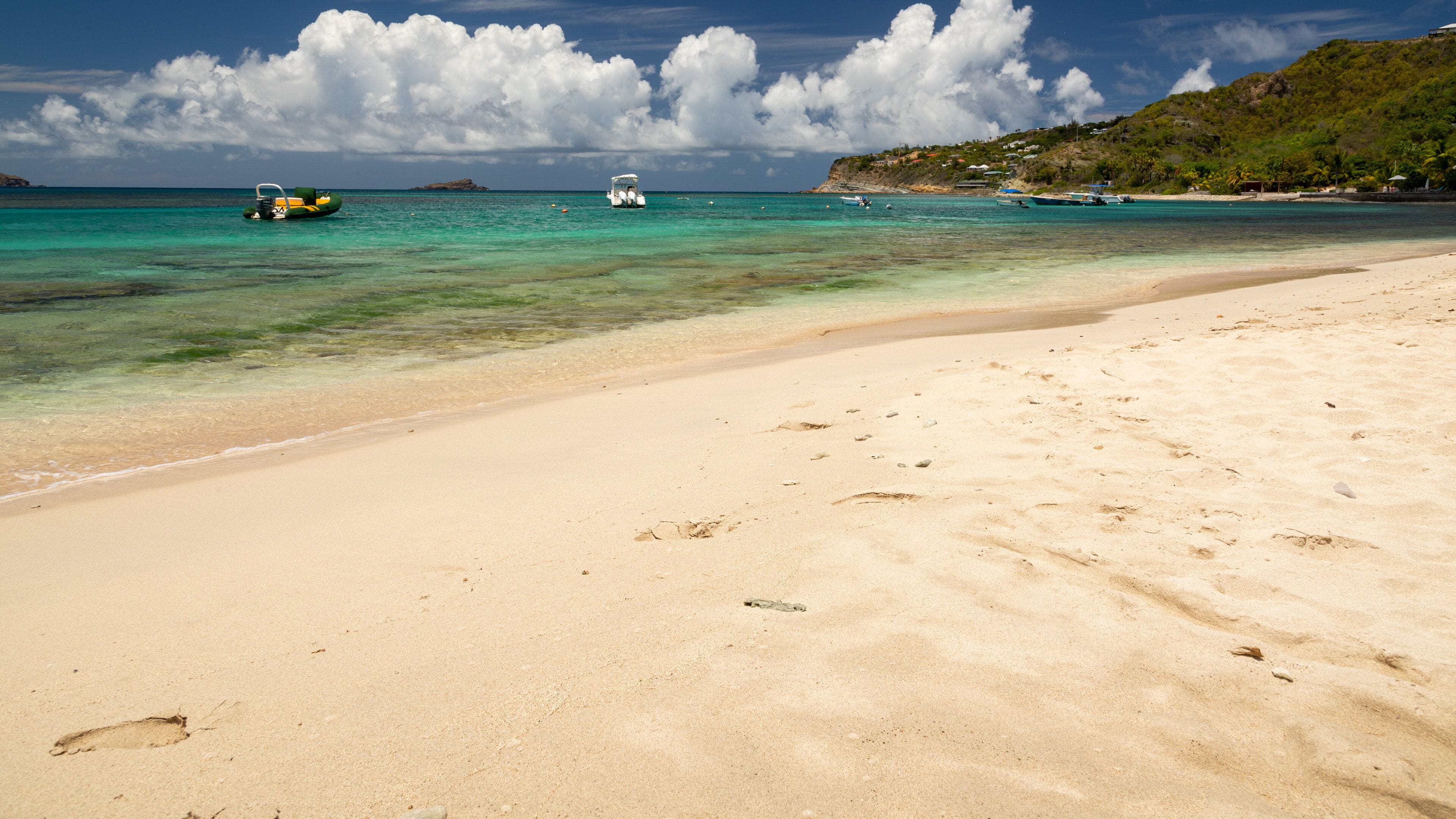 A sandy beach with footprints, clear turquoise water, and boats in the distance.