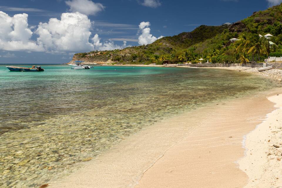 A clear turquoise beach with a boat in the water, a sandy shore, and a hill with vegetation and buildings in the background.
