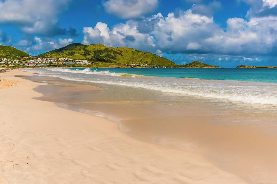 A beach with clear turquoise water, a sandy shore, and green hills in the background.