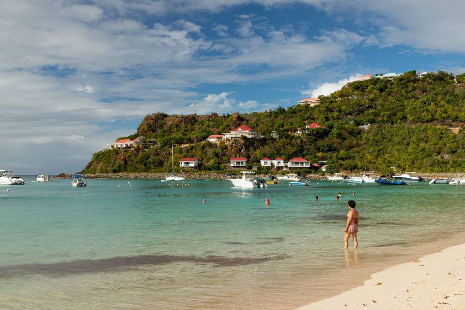 A beach with clear turquoise water, a sandy shore, and houses on a hillside.