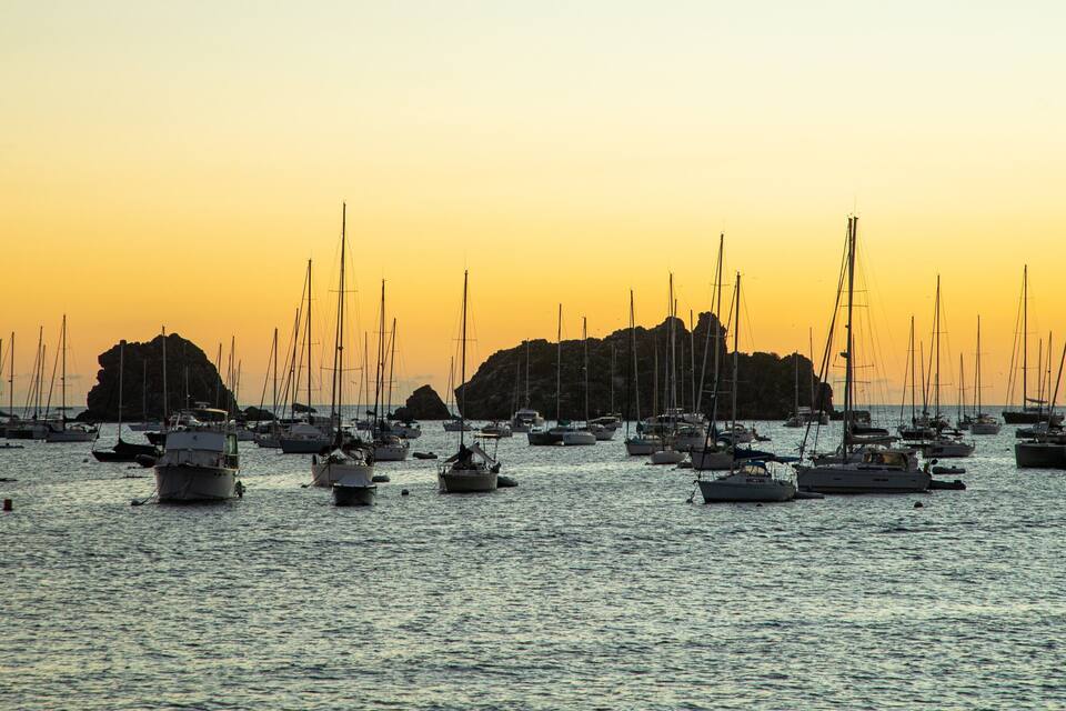 A marina with numerous sailboats and yachts anchored near rocky outcrops during sunset.
