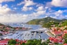 A coastal town with a marina filled with boats, surrounded by lush green hills and red-roofed buildings.