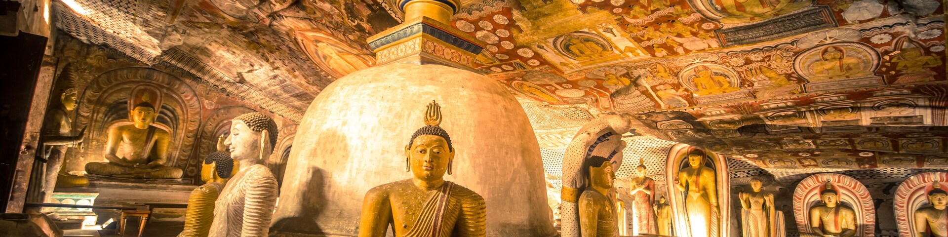 A temple interior with large Buddha statues, intricate ceiling artwork, and a central stupa.
