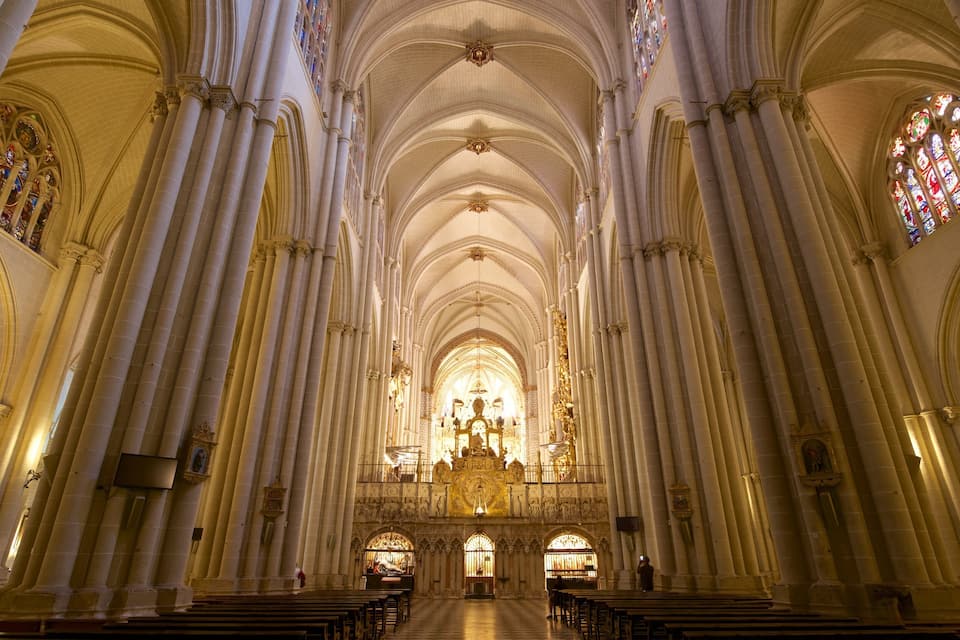 A cathedral interior with high vaulted ceilings, large stained glass windows, and rows of pews.
