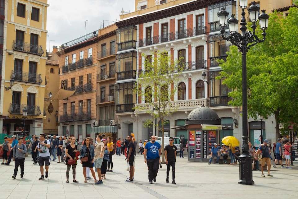 A bustling pedestrian area with historic buildings, a prominent yellow umbrella, and a central tree.