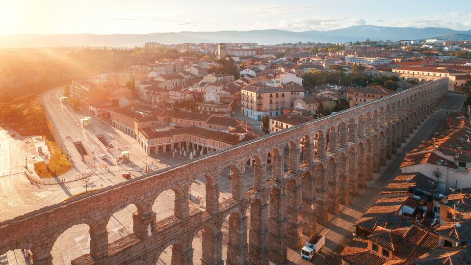 A historic stone bridge with arches, a cityscape with buildings, and a mountain in the background.