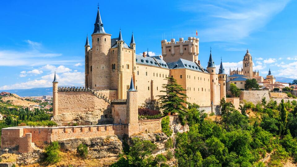 A historic castle with multiple towers and a fortified wall, surrounded by greenery and a clear sky.
