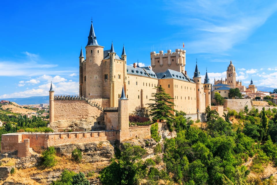 A historic castle with multiple towers and a fortified wall, surrounded by greenery and a clear sky.