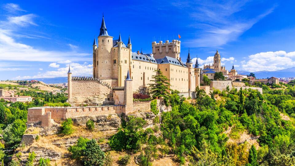 A historic castle with multiple towers and a surrounding landscape of trees and hills.