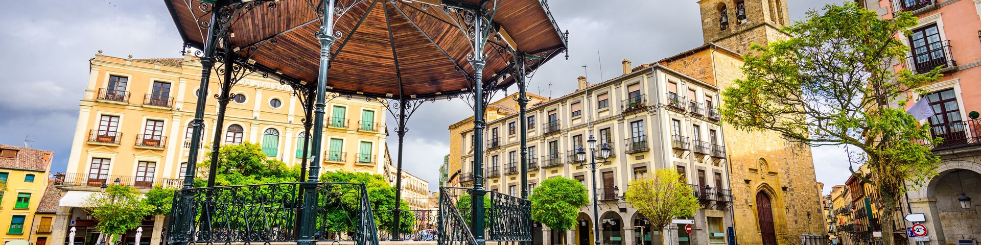 Un pabellón cubierto con intrincados detalles metálicos, una plaza adoquinada y edificios históricos con una torre destacada.