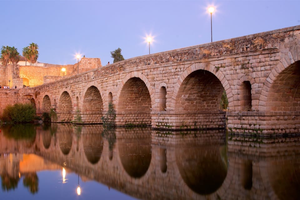 A stone bridge with multiple arches over a calm river, reflecting the bridge and surrounding trees.