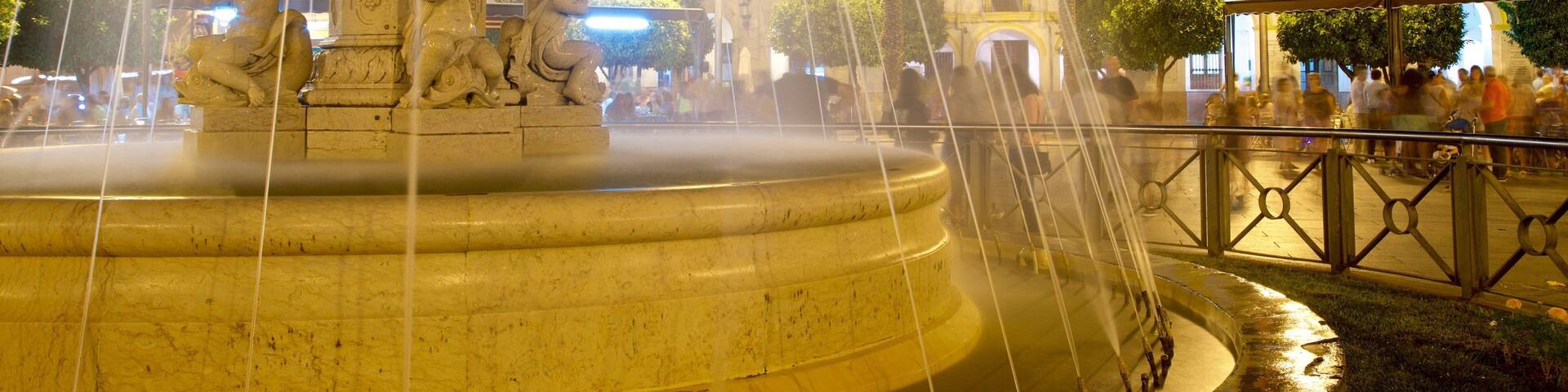 A nighttime fountain with water jets, a clock tower, and a crowd of people in the background.