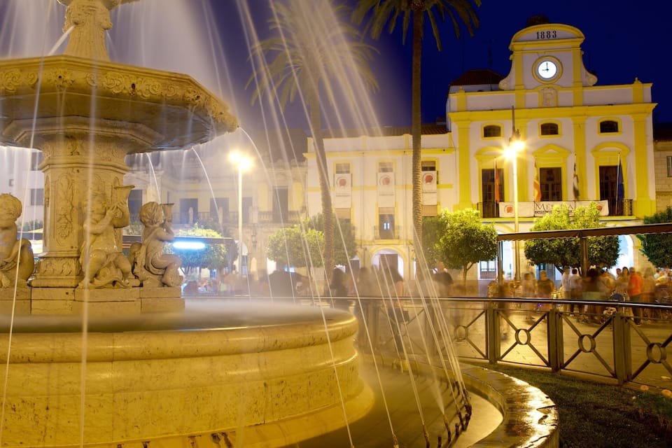 Una fuente nocturna con chorros de agua, una torre del reloj y una multitud de personas al fondo.