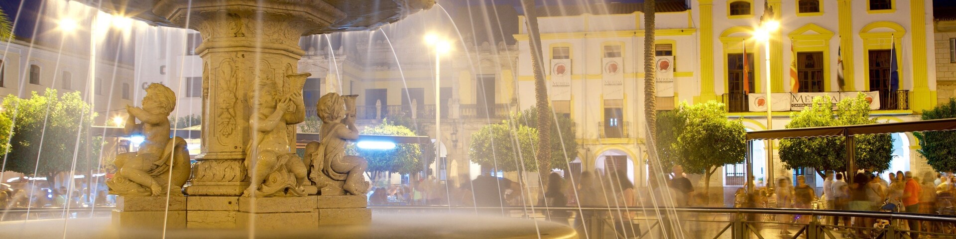Una fuente nocturna con chorros de agua, una torre del reloj y una multitud de personas al fondo.