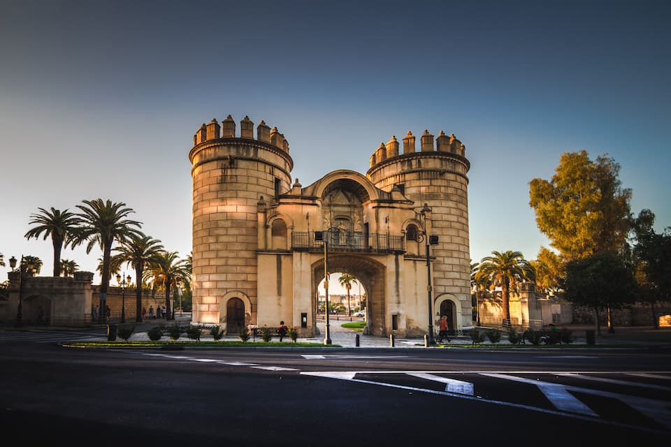 A historic stone archway with two towers, palm trees, and a clear sky.