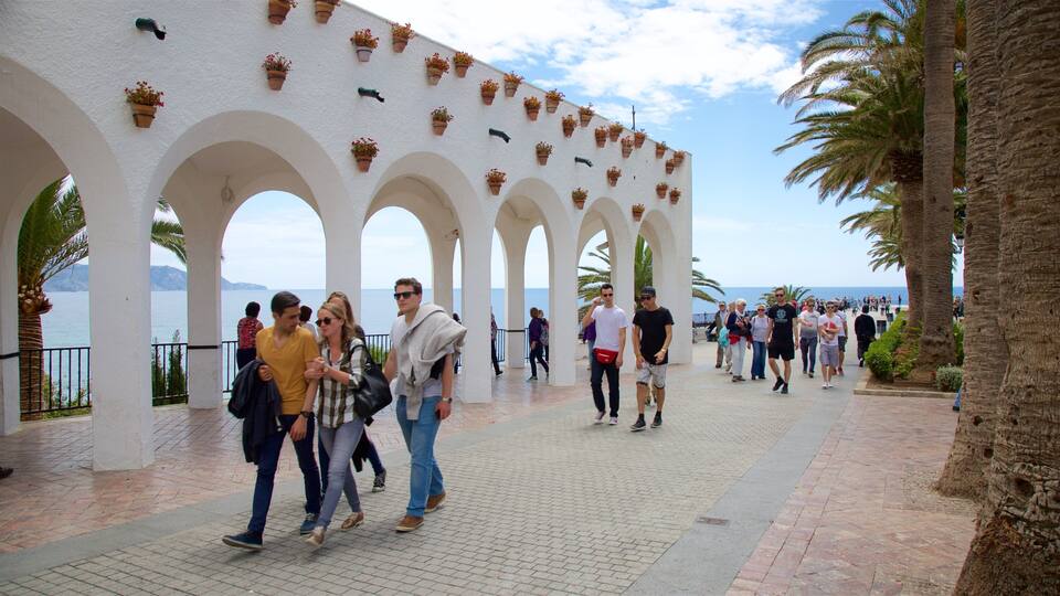 A coastal walkway with white arches and potted plants, leading to the sea.