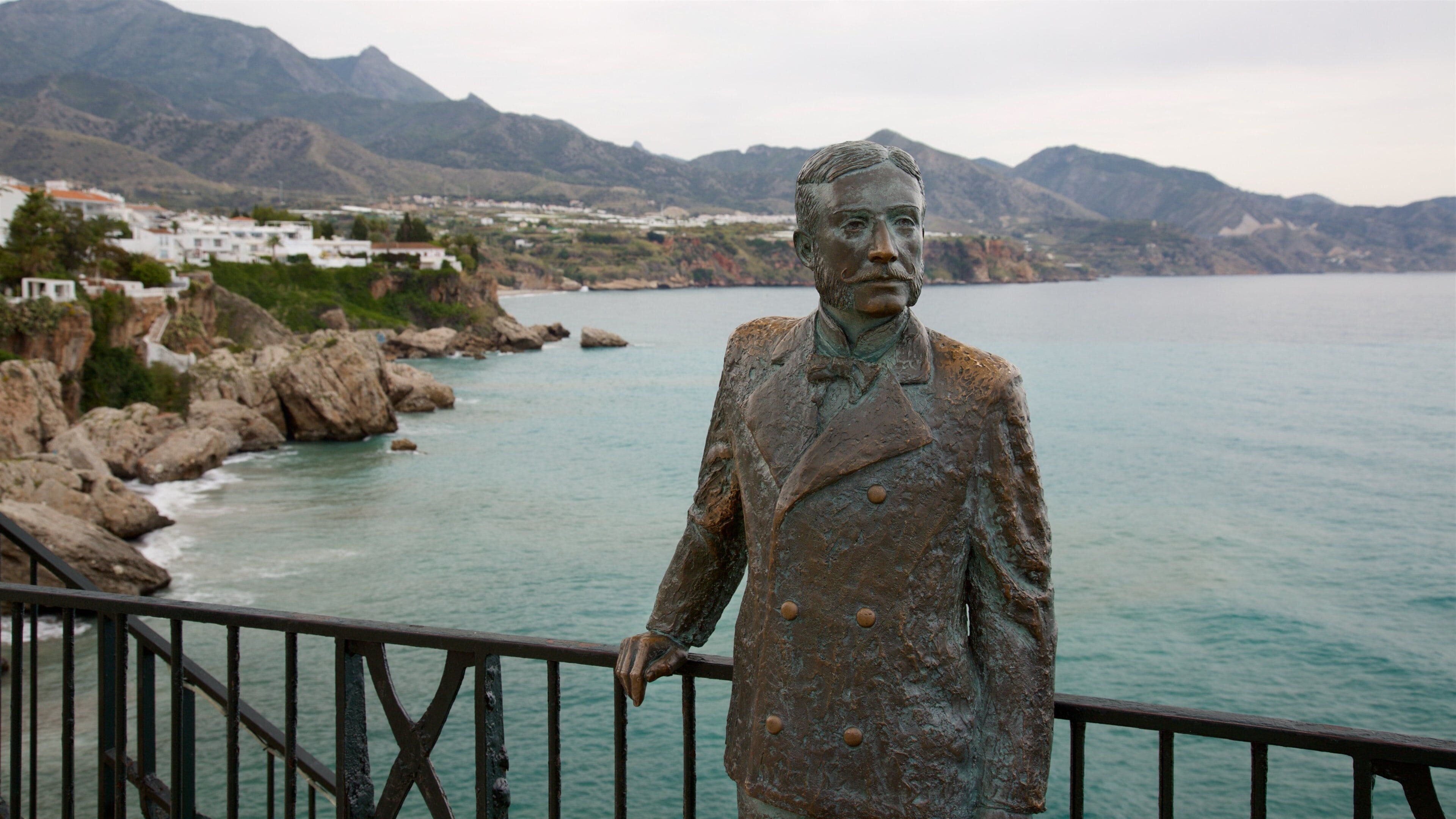 A statue of a man in a suit by a coastal area with rocky cliffs and a view of a town in the background.