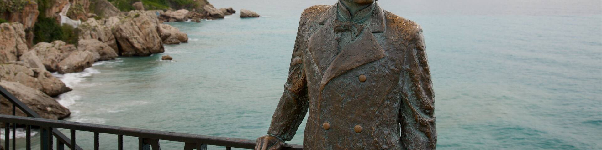A statue of a man in a suit by a coastal area with rocky cliffs and a view of a town in the background.