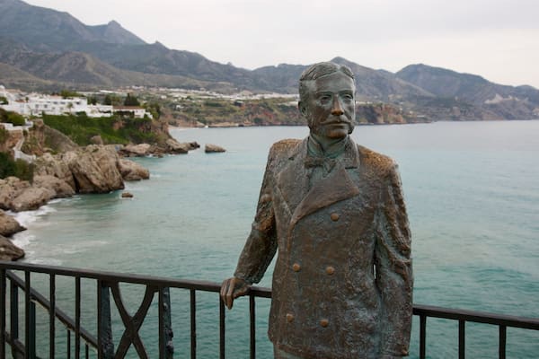 Une statue d’un homme en costume, située près d’une zone côtière avec des falaises rocheuses, et en arrière-plan, on aperçoit une ville.